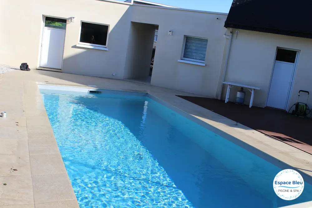 Piscine à eau bleue devant une maison, margelle beige